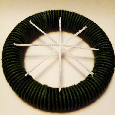 Four white crochet chains attached radially to a green yarn-wrapped foam ring, forming a cross